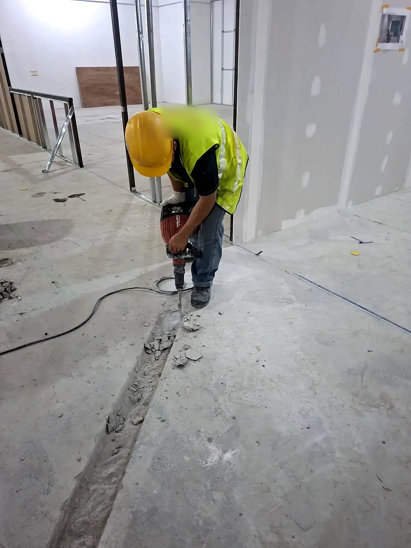 Electrician in high-visibility vest and hard hat chasing a floor slot for conduit with a breaker.