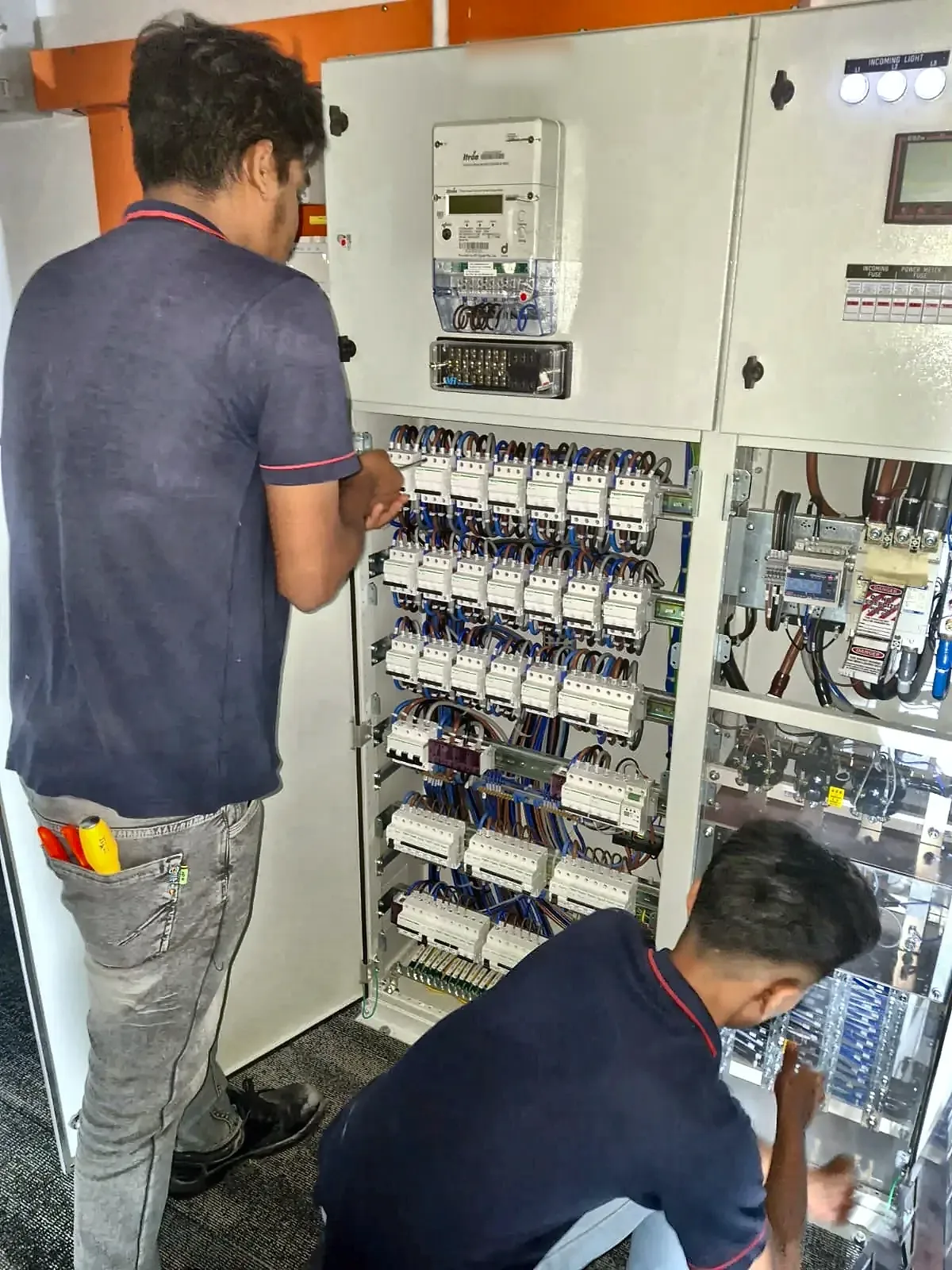 Two electricians commissioning an LV panel, working across meters and rows of miniature circuit breakers.
