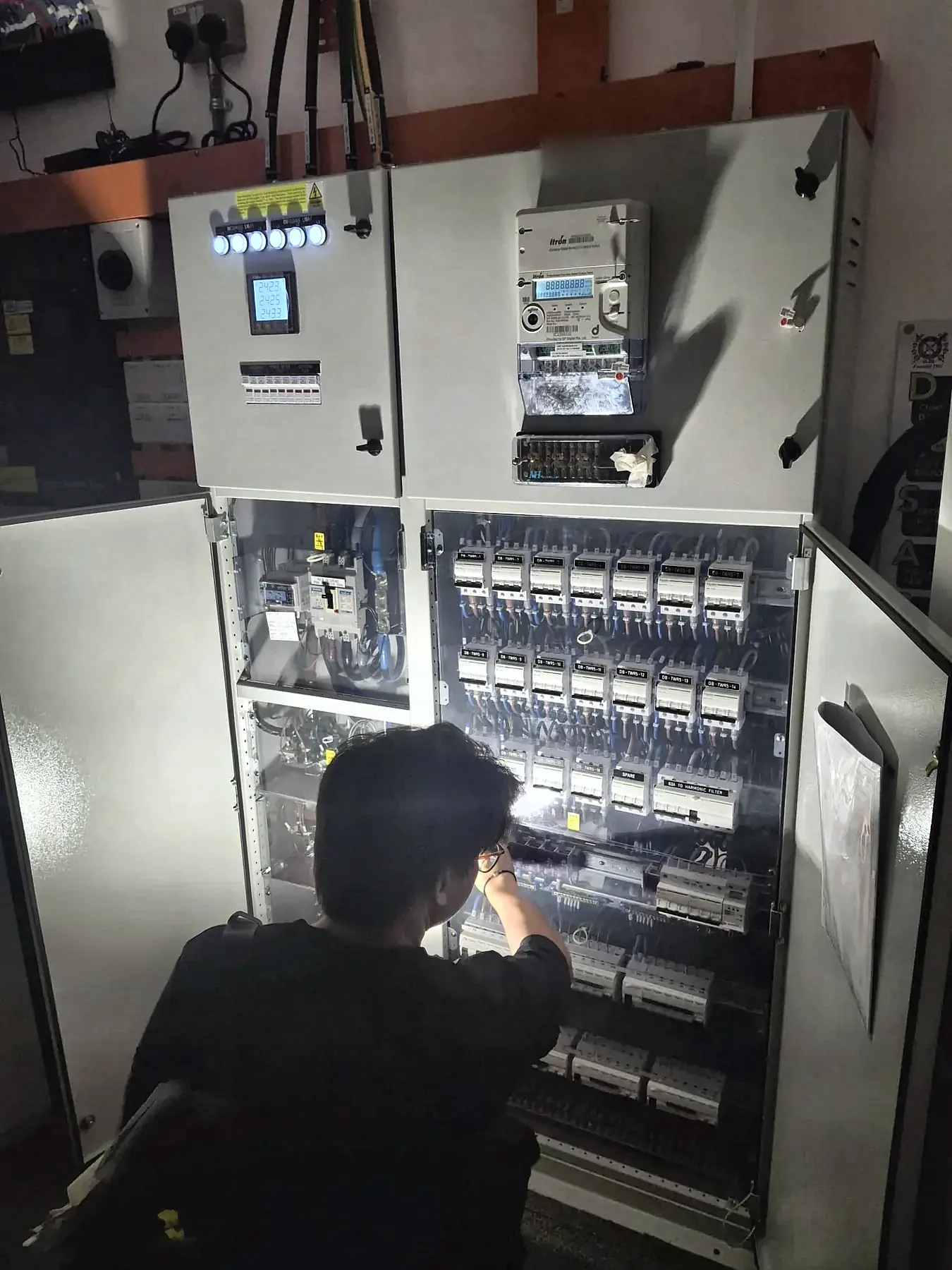 Electrician testing inside an open switchboard with controller and dense rows of circuit breakers.